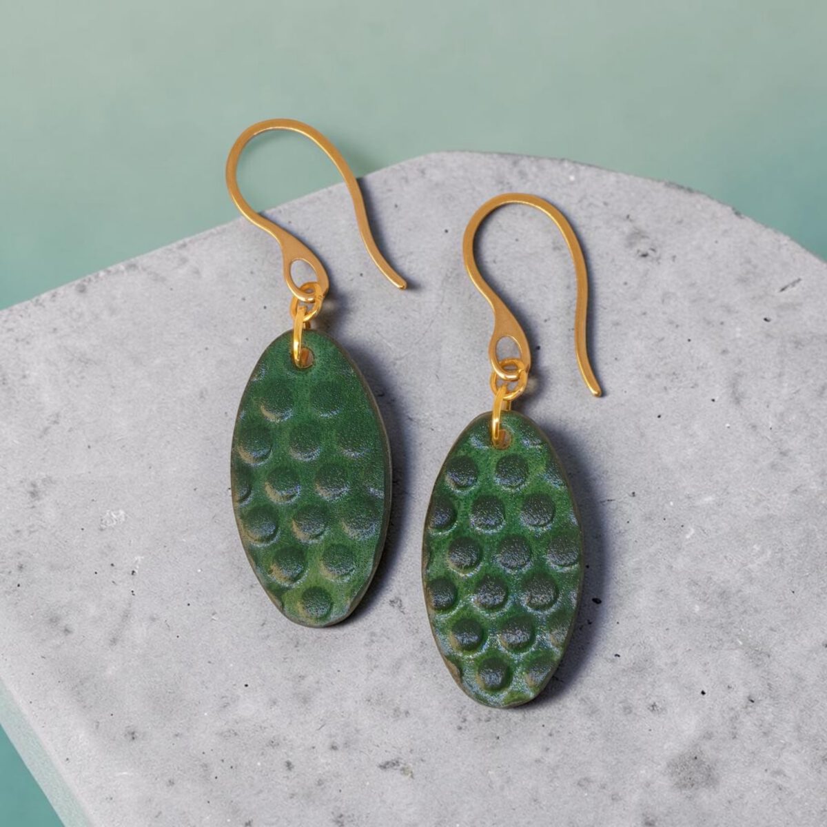 Boucles d'oreilles ovales en cuir teinte vert forêt, texturage ronds, vues de face, suspendues à des boucles dorées sur béton