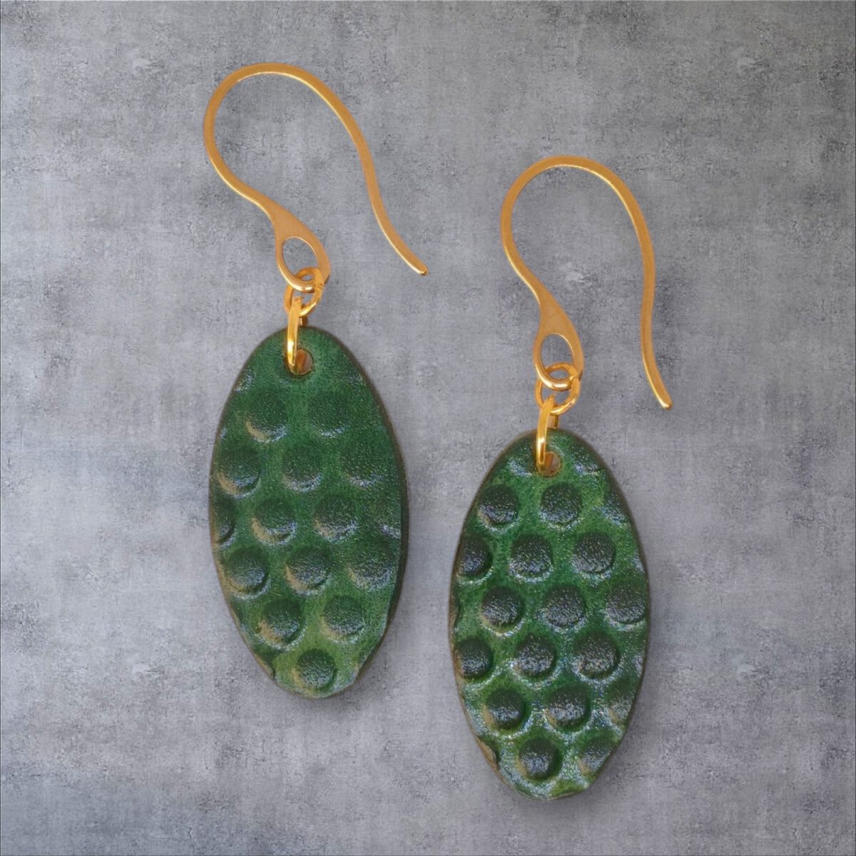 Boucles d'oreilles ovales en cuir teinte vert forêt, texturage ronds, vues de face, suspendues à des boucles dorées sur fond gris
