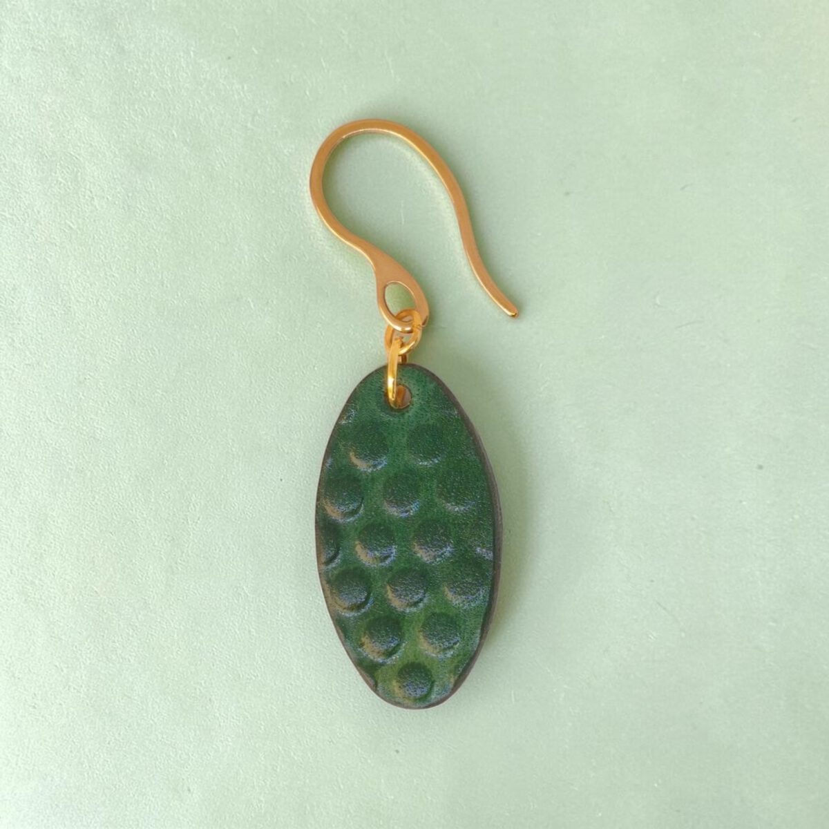 Boucle d'oreille ovale en cuir teinte vert forêt, texturage ronds, vue de face, suspendue à une boucle dorée, fond design