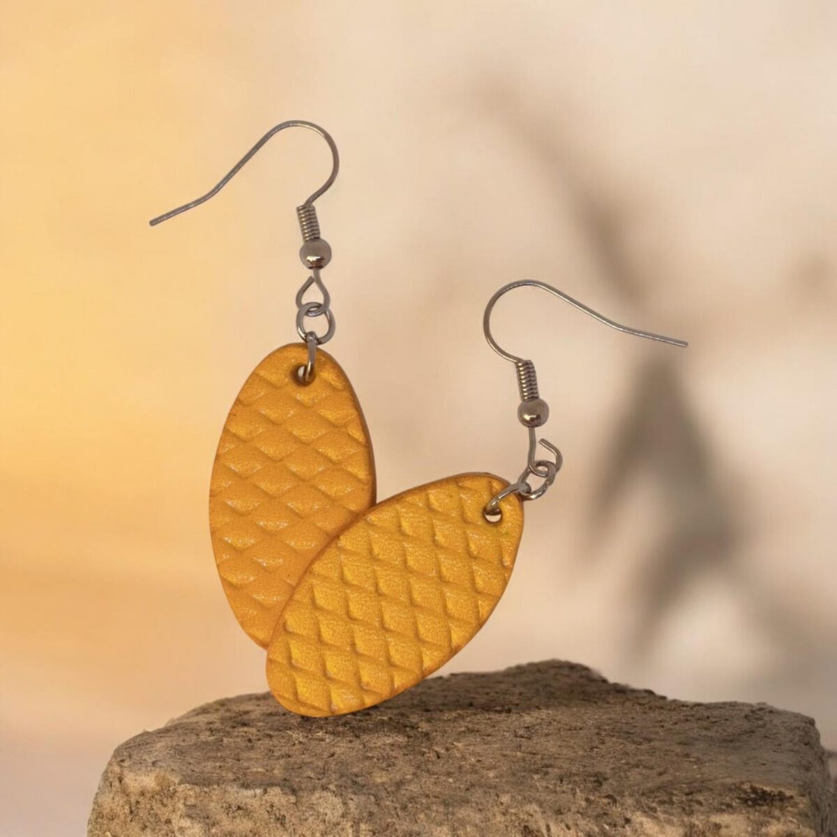 Boucles d'oreilles ovales en cuir teinte jaune, texturage poisson fin, vues de face se chevauchant, suspendues à des boucles argentées sur pierre
