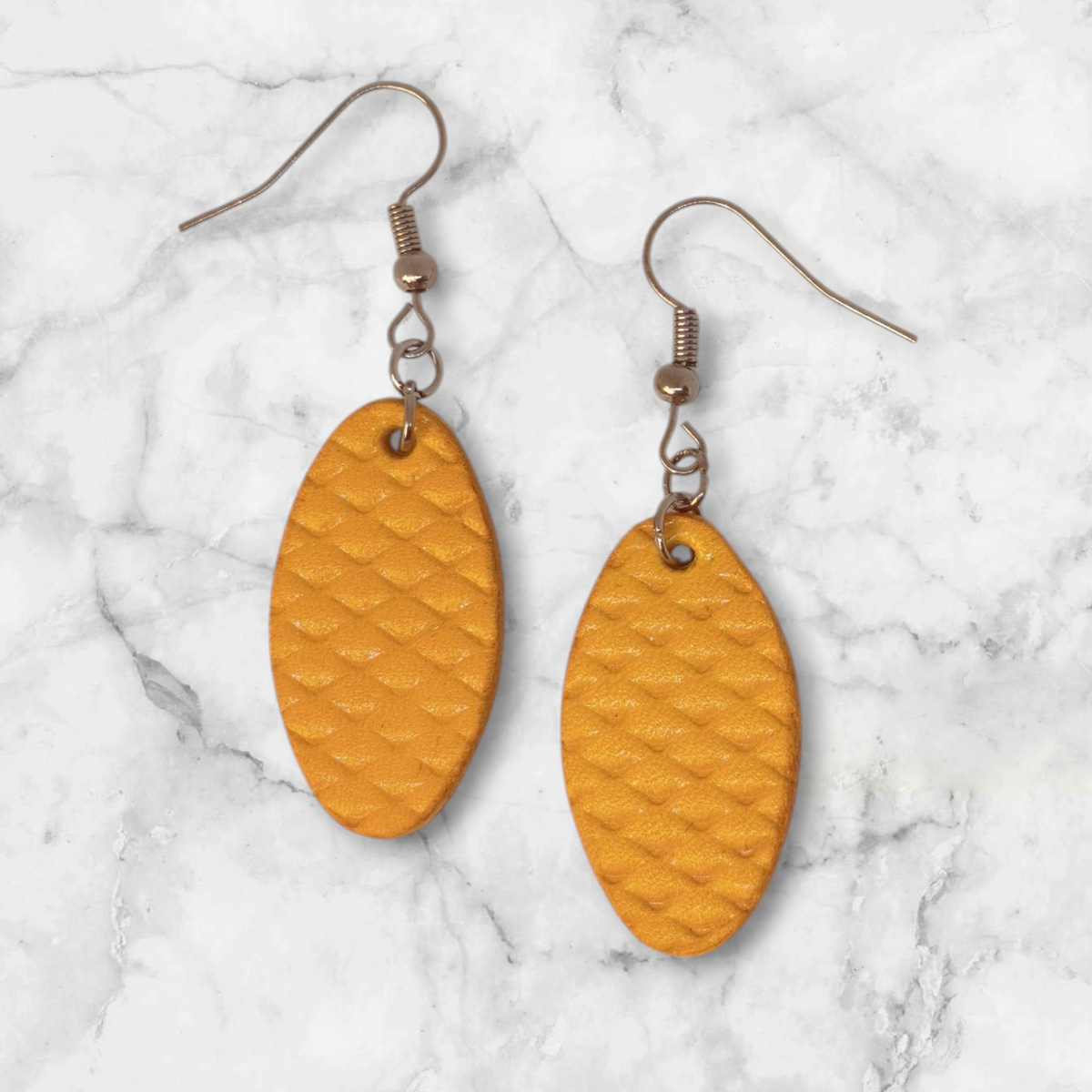 Boucles d'oreilles ovales en cuir teinte jaune, texturage poisson fin, vues de face, suspendues à des boucles argentées sur marbre