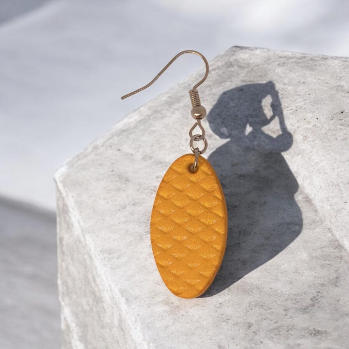 Boucle d'oreille ovale en cuir teinte jaune, texturage poisson fin, vue de face, suspendue à une boucle argentée, sur pierre