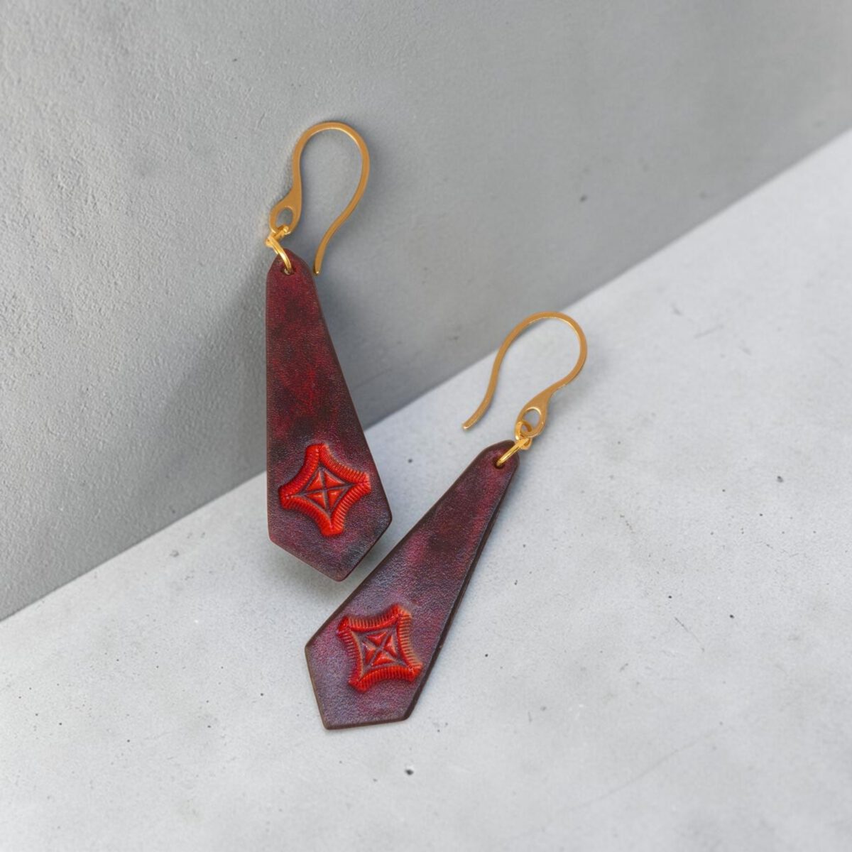 Boucles d'oreilles en cuir teinte acajou, estampage croix teinte rouge, vues de face, suspendues à des boucles dorées sur béton