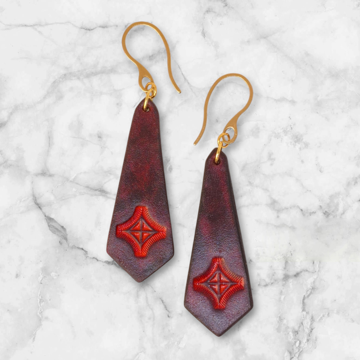 Boucles d'oreilles en cuir teinte acajou, estampage croix teinte rouge, vues de face, suspendues à des boucles dorées posé sur du marbre