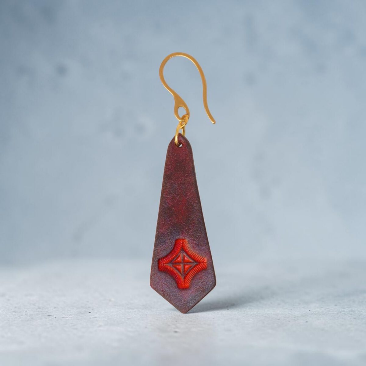 Boucle d'oreille en cuir teinte acajou, estampage croix rouge, vue de face, suspendue à une boucle dorée, fond design