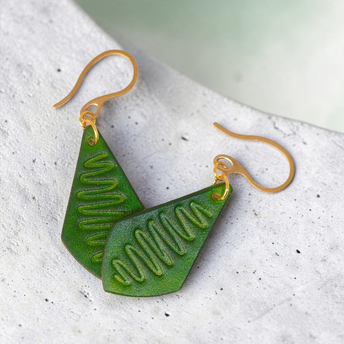Boucles d'oreilles forme larme en cuir teinture vert forêt, estampage "Lombard Street", vues de face se chevauchant, suspendues à des boucles dorées sur béton