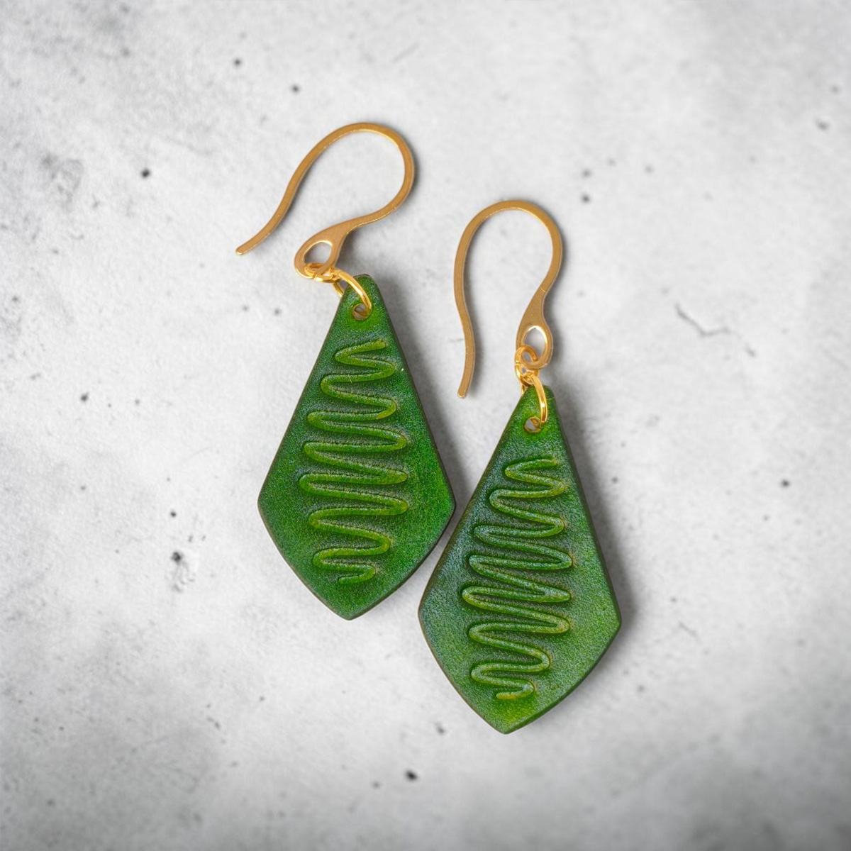Boucles d'oreilles forme larme en cuir teinture vert forêt, estampage "Lombard Street", vues de face, suspendues à des boucles dorées sur béton