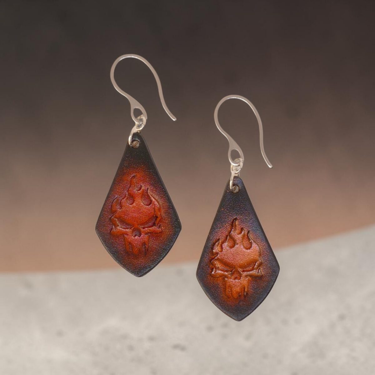 Boucles d'oreilles forme larme en cuir teinture noire, estampage tête de mort couleur cognac en dégradé, vues de face, suspendues à des boucles argentées, fond design