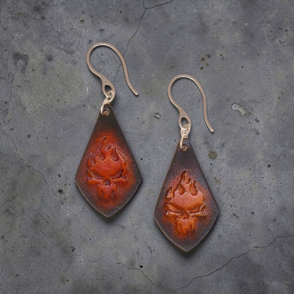 Boucles d'oreilles forme larme en cuir teinture noire, estampage tête de mort couleur cognac en dégradé, vues de face, suspendues à des boucles argentées sur béton craquelé