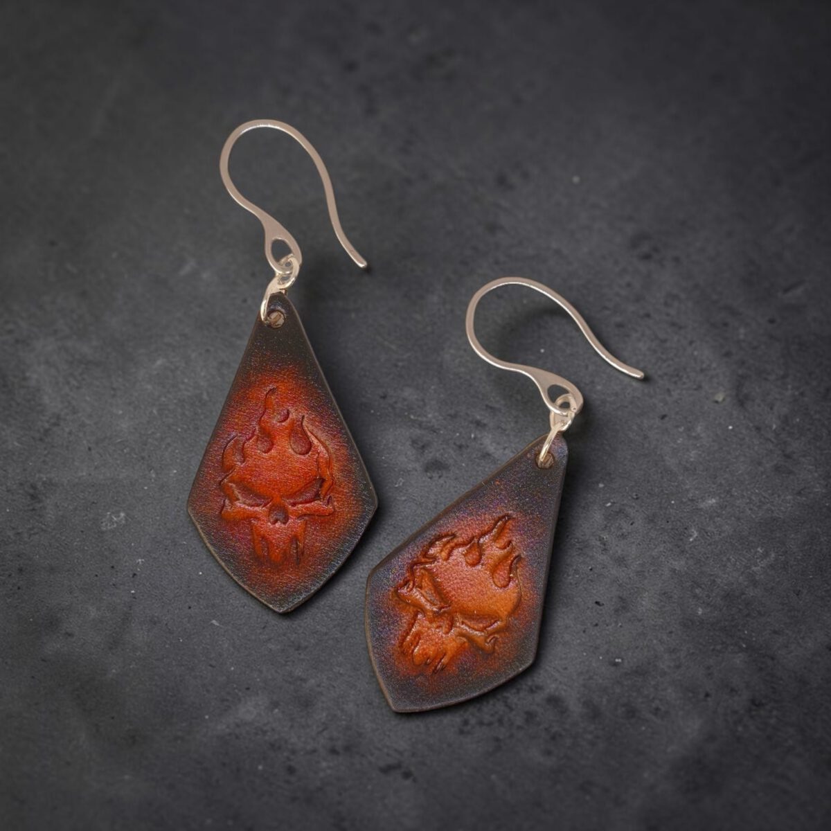 Boucles d'oreilles forme larme en cuir teinture noire, estampage tête de mort couleur cognac en dégradé, vues de face, suspendues à des boucles argentées sur fond noir
