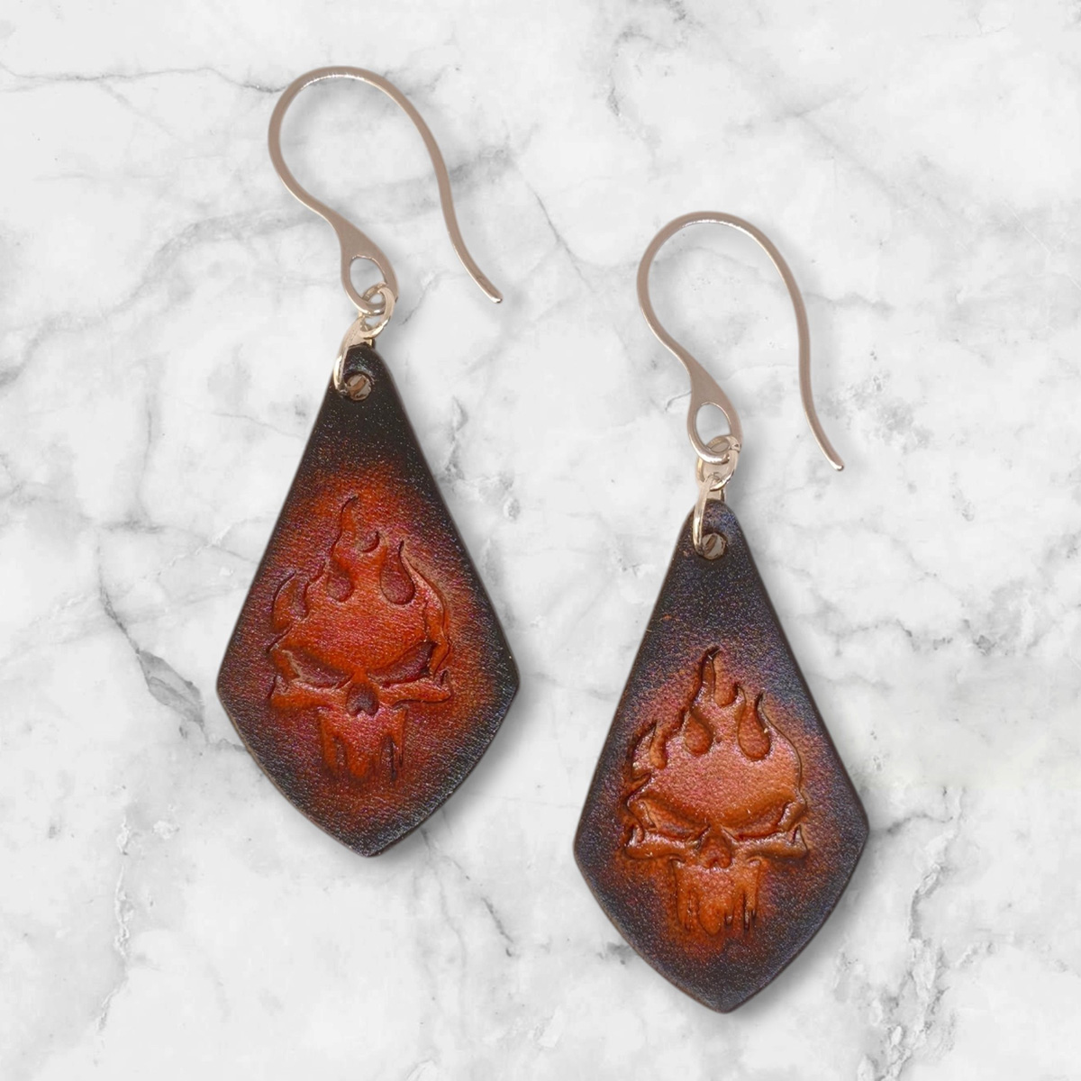 Boucles d'oreilles forme larme en cuir teinture noire, estampage tête de mort couleur cognac en dégradé, vues de face, suspendues à des boucles argentées sur marbre