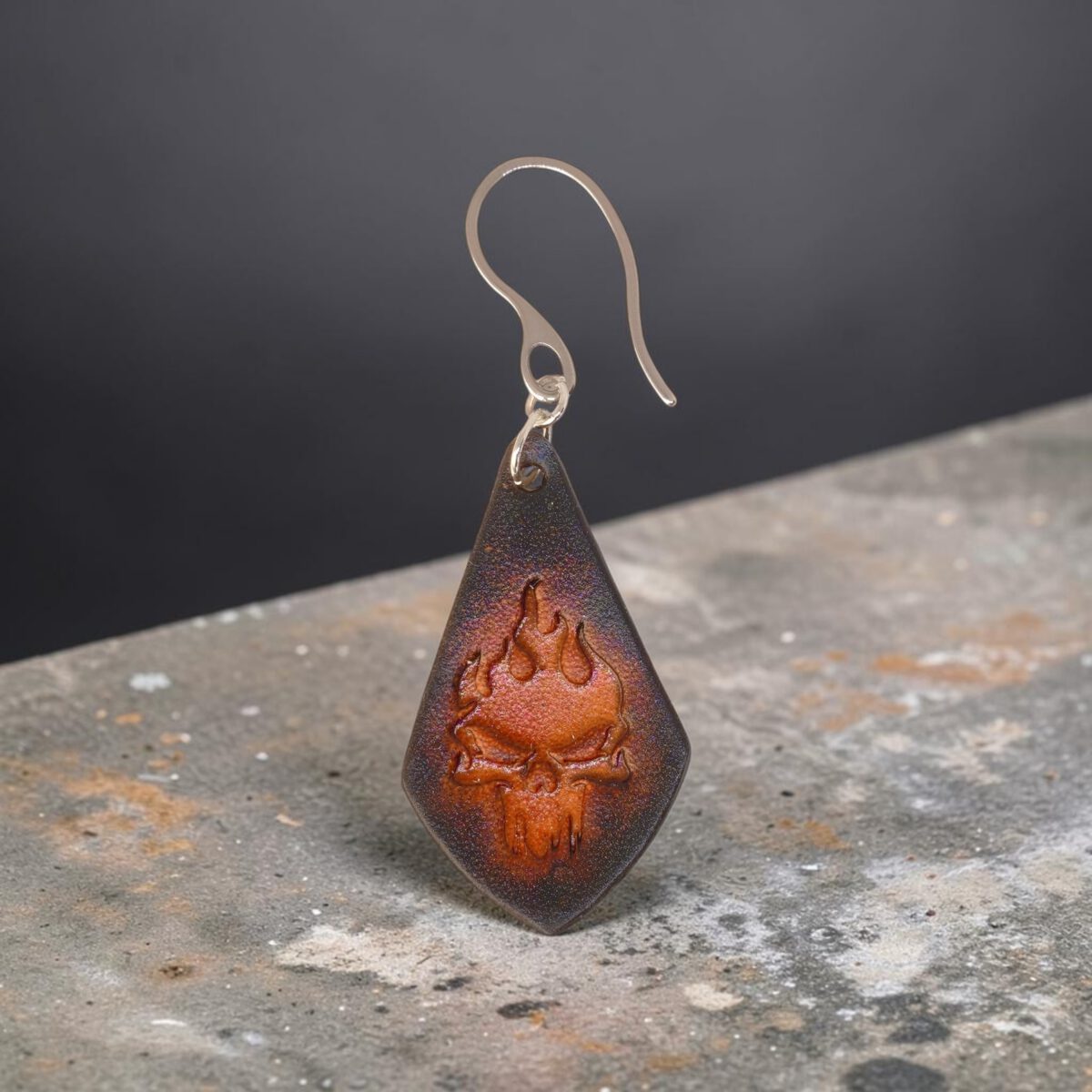 Boucle d'oreille forme larme en cuir teinture noire, estampage tête de mort couleur cognac en dégradé, vue de face, suspendue à une boucle argentée, sur pierre