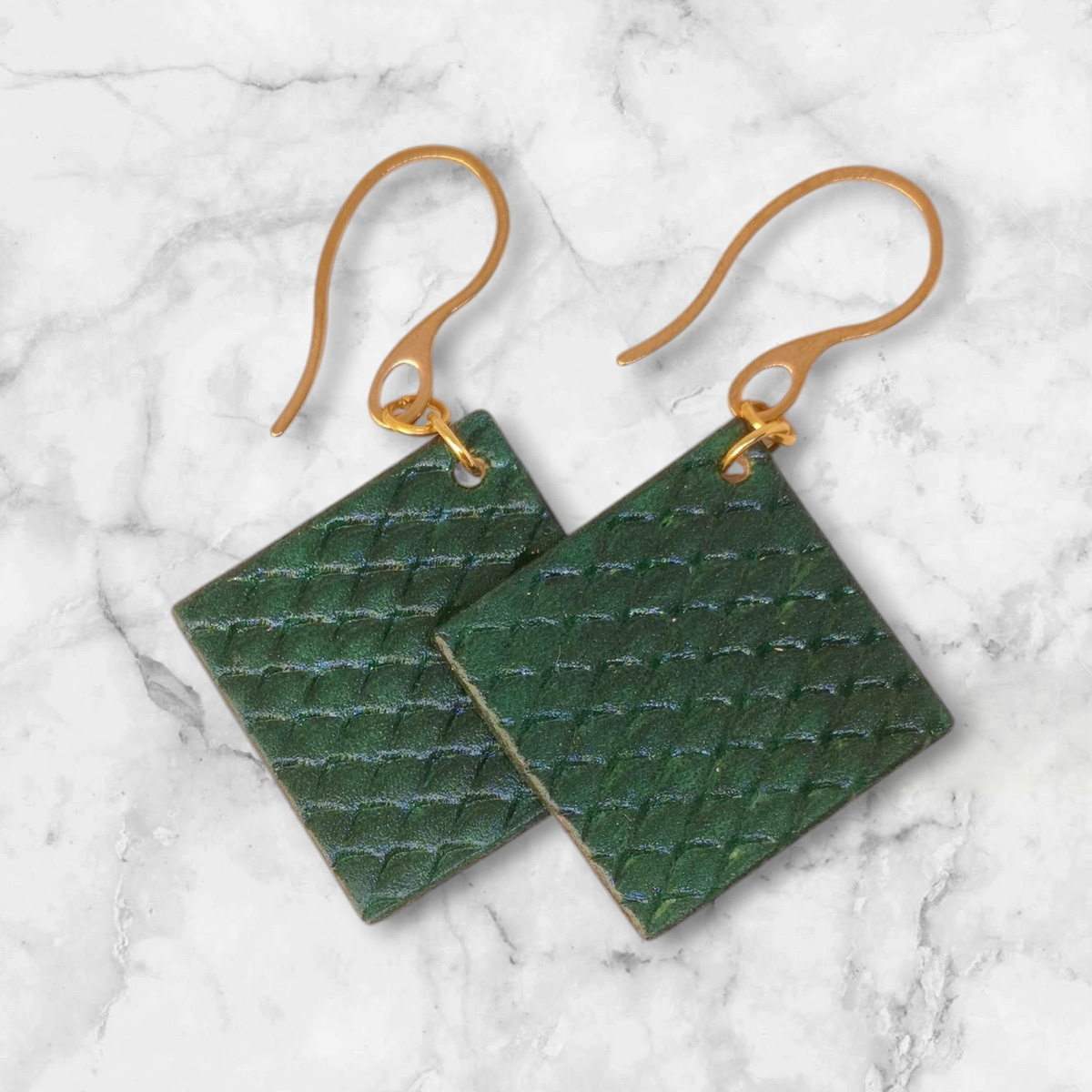 Boucles d'oreilles carrées en cuir teinté vert foncé, texturage poisson, vues de face se chevauchant, suspendues à des boucles dorées sur marbre