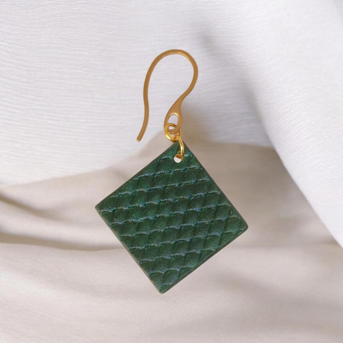 Boucle d'oreille carrée en cuir teinté vert foncé, texturage poisson, vue de face, suspendue à une boucle dorée, fond tissu