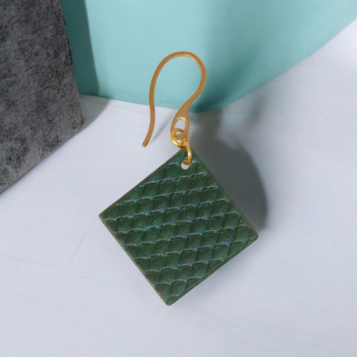 Boucle d'oreille carrée en cuir teinté vert foncé, texturage poisson, vue de face, suspendue à une boucle dorée, fond design