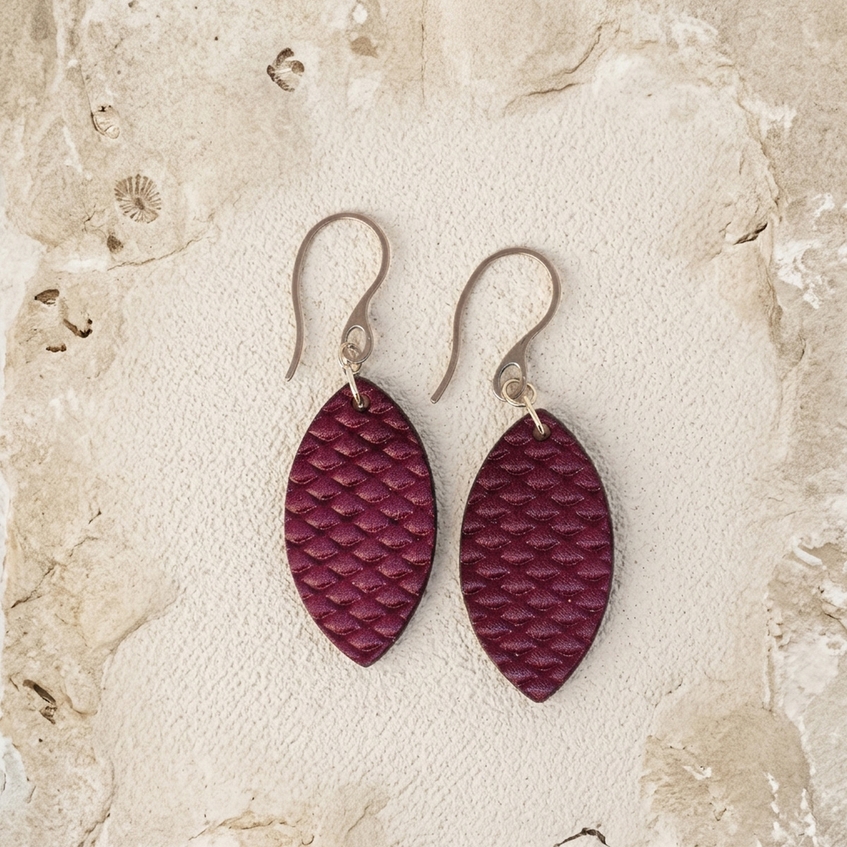 Boucles d'oreilles forme poisson en cuir teinture violet, texturage poisson fin, vues de face, suspendues à des boucles argentées sur sable