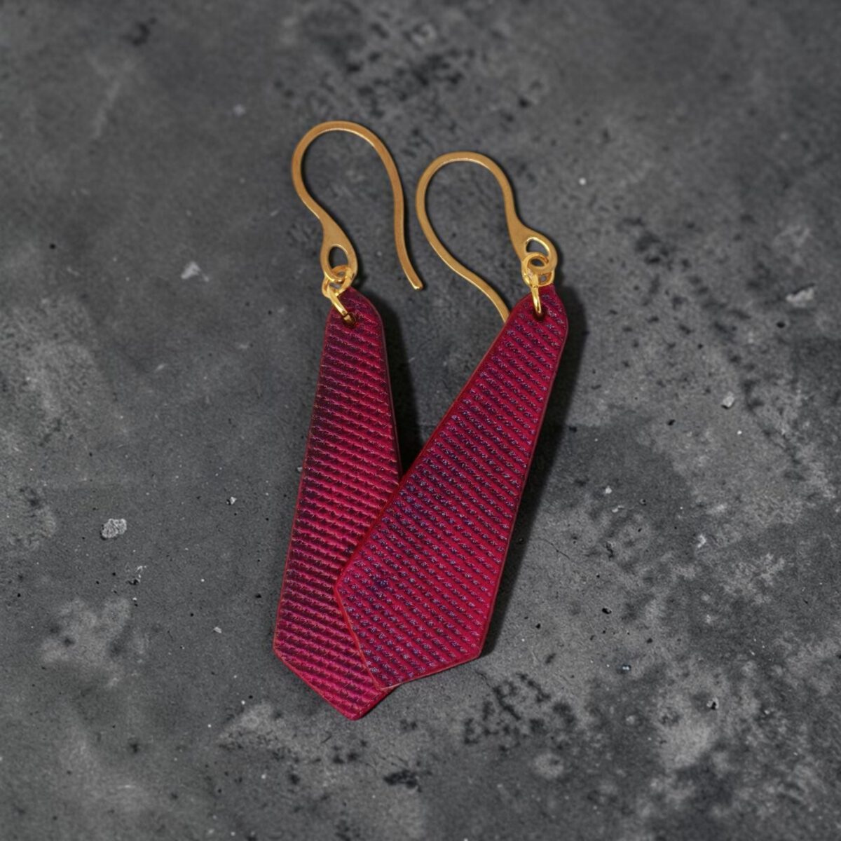 Boucles d'oreilles cravates en cuir teinté fuschia, finition lime vieillie, vues de face se chevauchant, suspendues à des boucles dorées sur un mannequin virtuel, sur béton