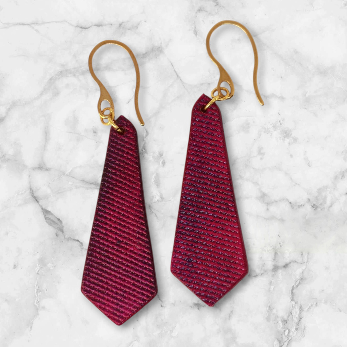 Boucles d'oreilles cravates en cuir teinté fuschia, finition lime vieillie, vues de face, suspendues à des boucles dorées sur marbre