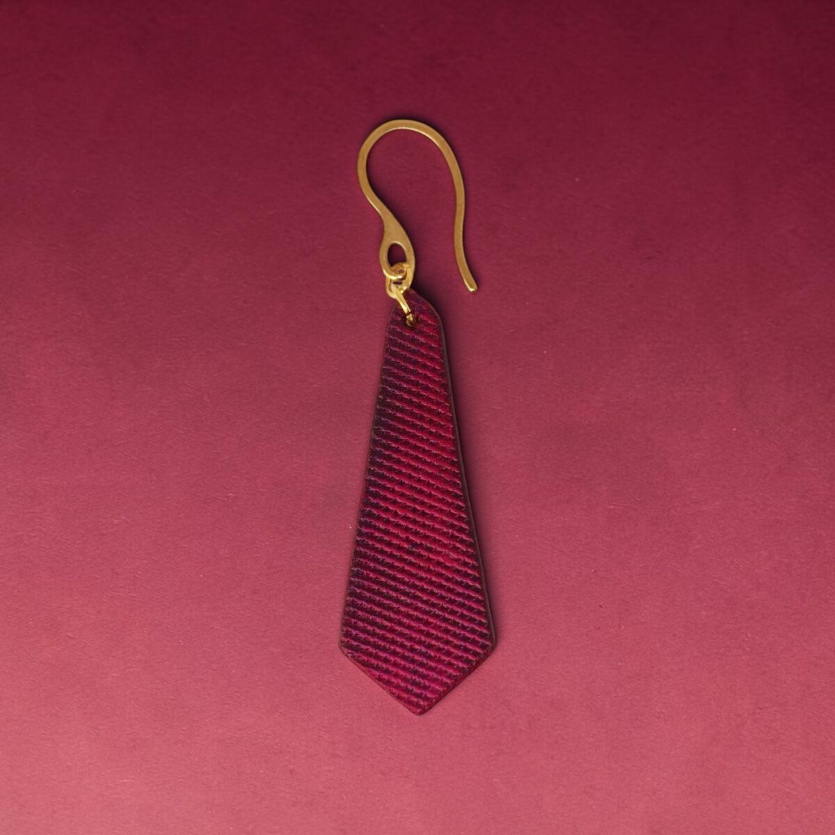 Boucle d'oreille cravate en cuir teinté fuschia, finition lime vieillie, vue de face, suspendue à une boucle dorée, fond design