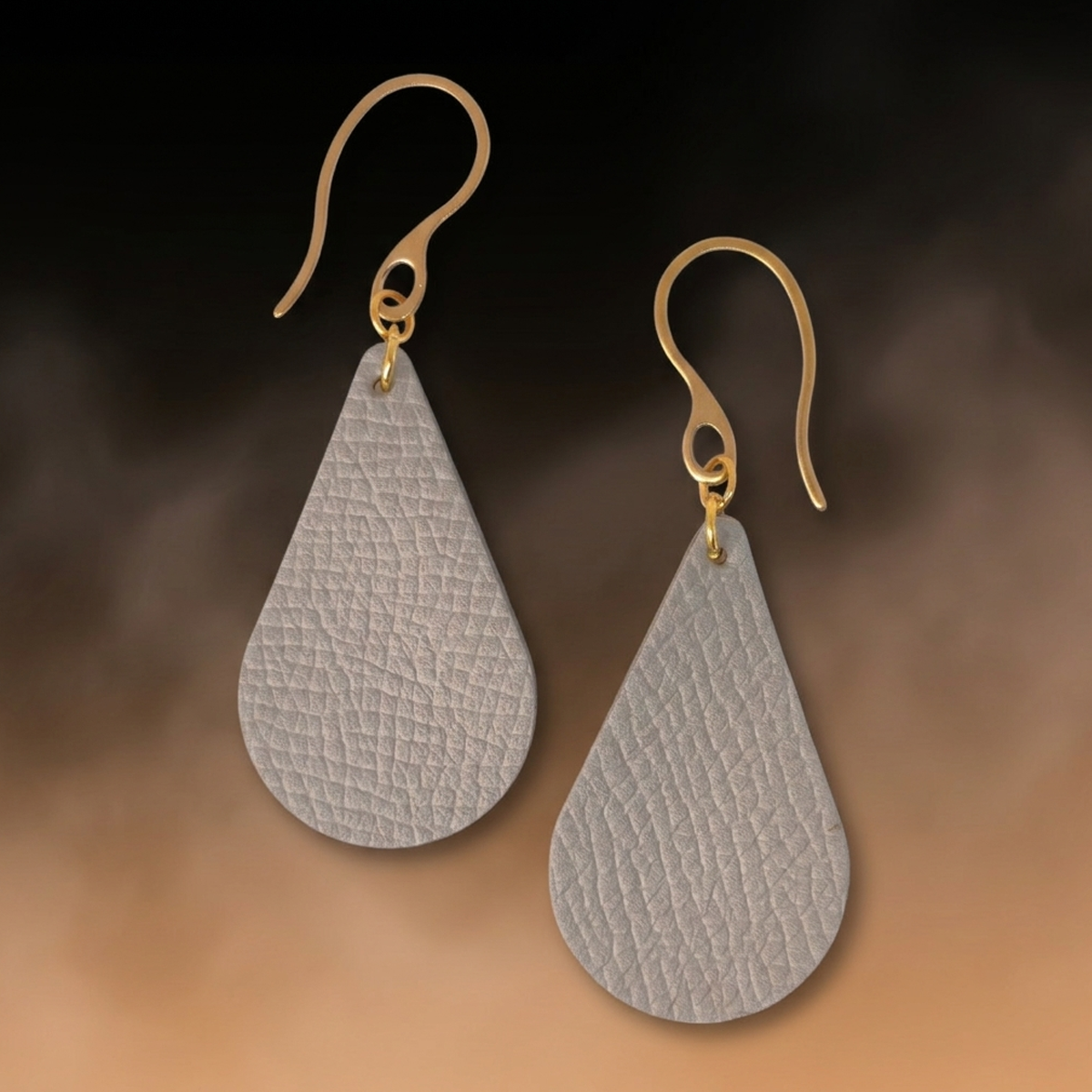 Boucles d'oreilles forme goutte d'eau en cuir gris, texture grainée, vues de face, suspendues à des boucles dorées sur fond dégradé