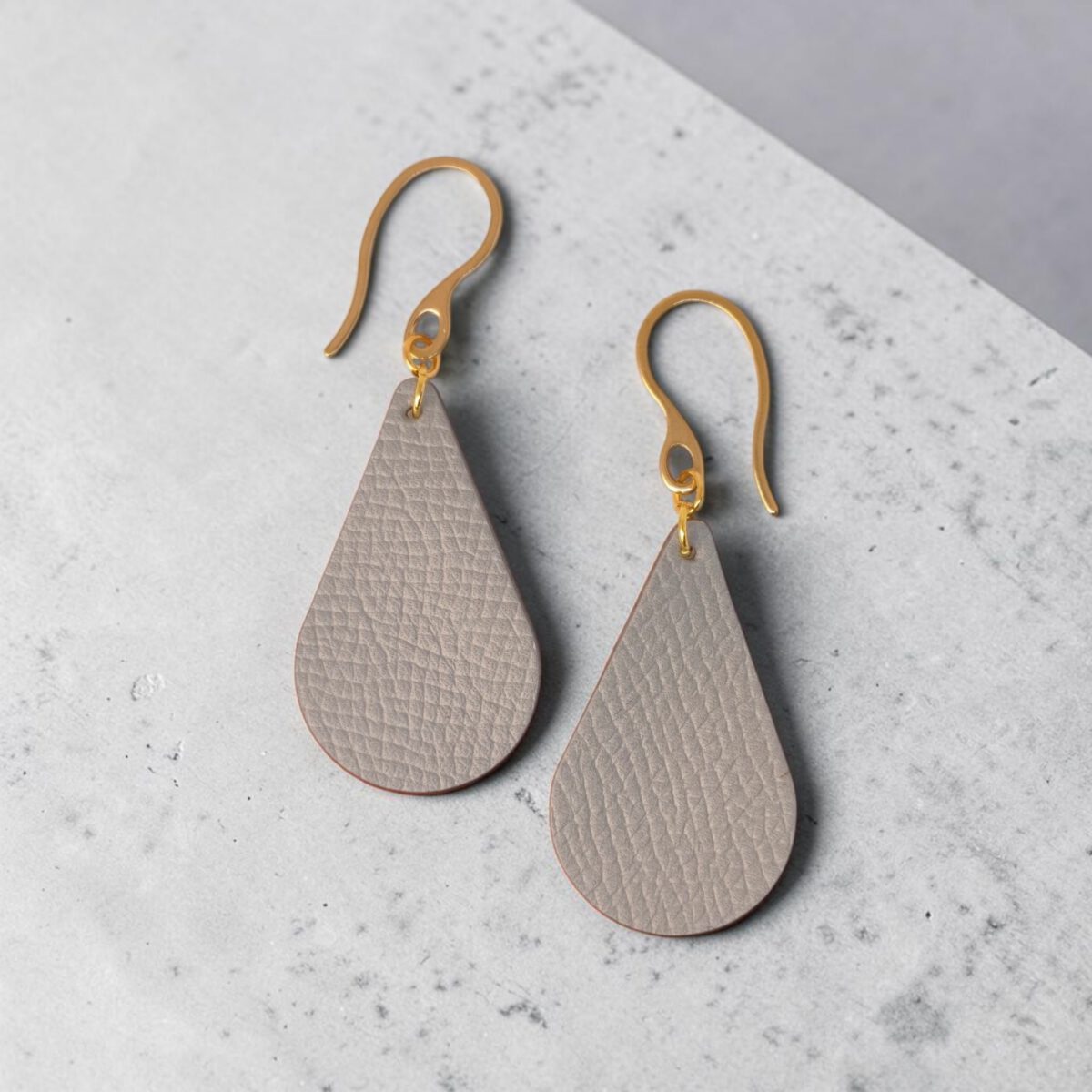 Boucles d'oreilles forme goutte d'eau en cuir gris, texture grainée, vues de face, suspendues à des boucles dorées sur béton