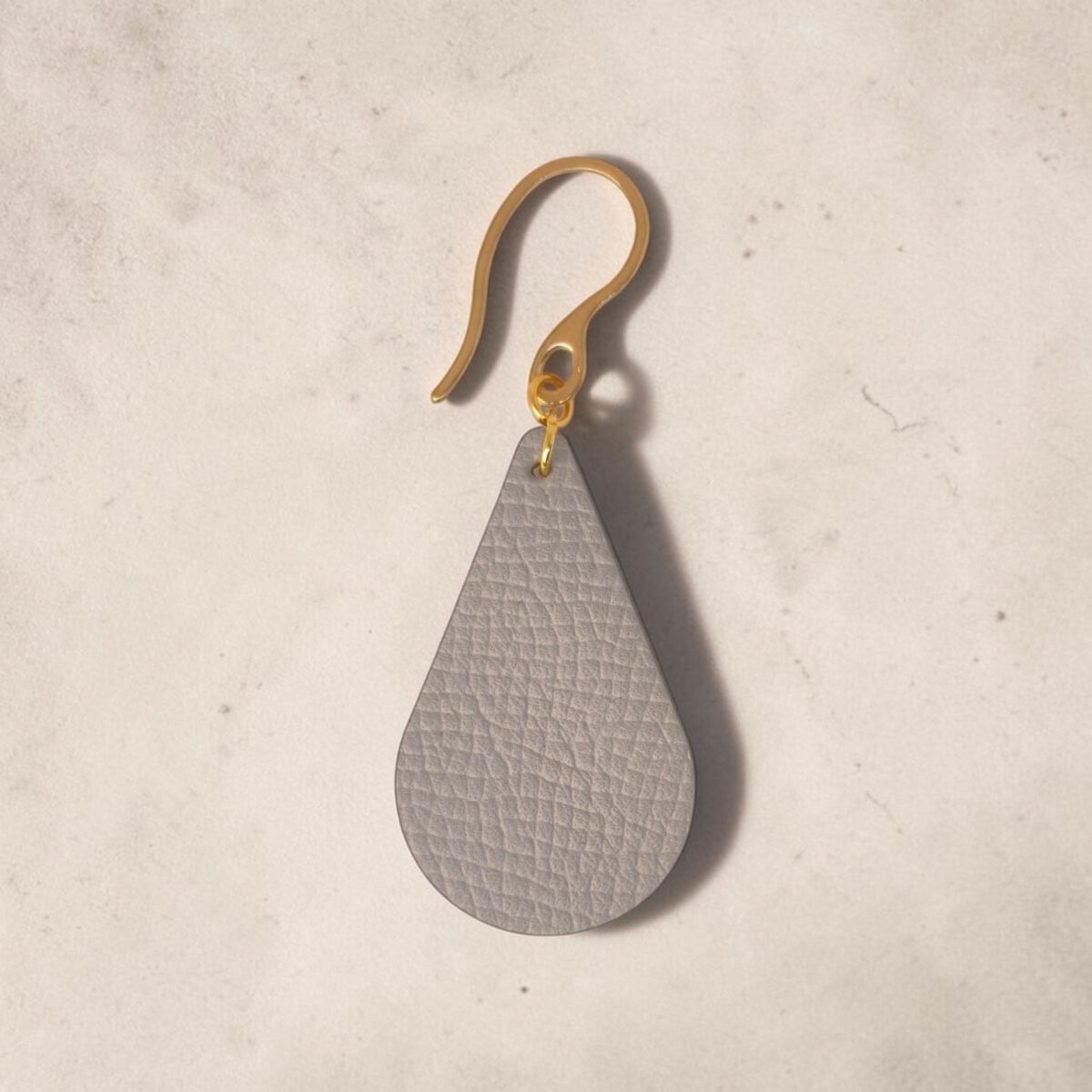 Boucle d'oreille forme goutte d'eau en cuir gris, texture grainée, vue de face, suspendue à une boucle dorée, fond design
