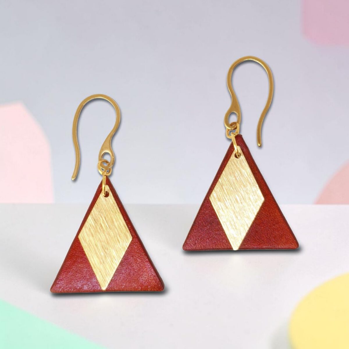 Boucles d'oreilles forme triangulaire en cuir teinture cognac, finition lisse et pendants losange dorés brossés vues de face, suspendues à des boucles dorées sur fond design