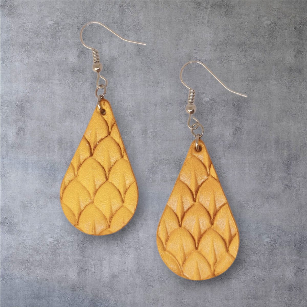 Boucles d'oreilles forme goutte d'eau en cuir teinture jaune, estampages au matoir écailles de poisson, vues de face, suspendues à des boucles argentées sur fond gris