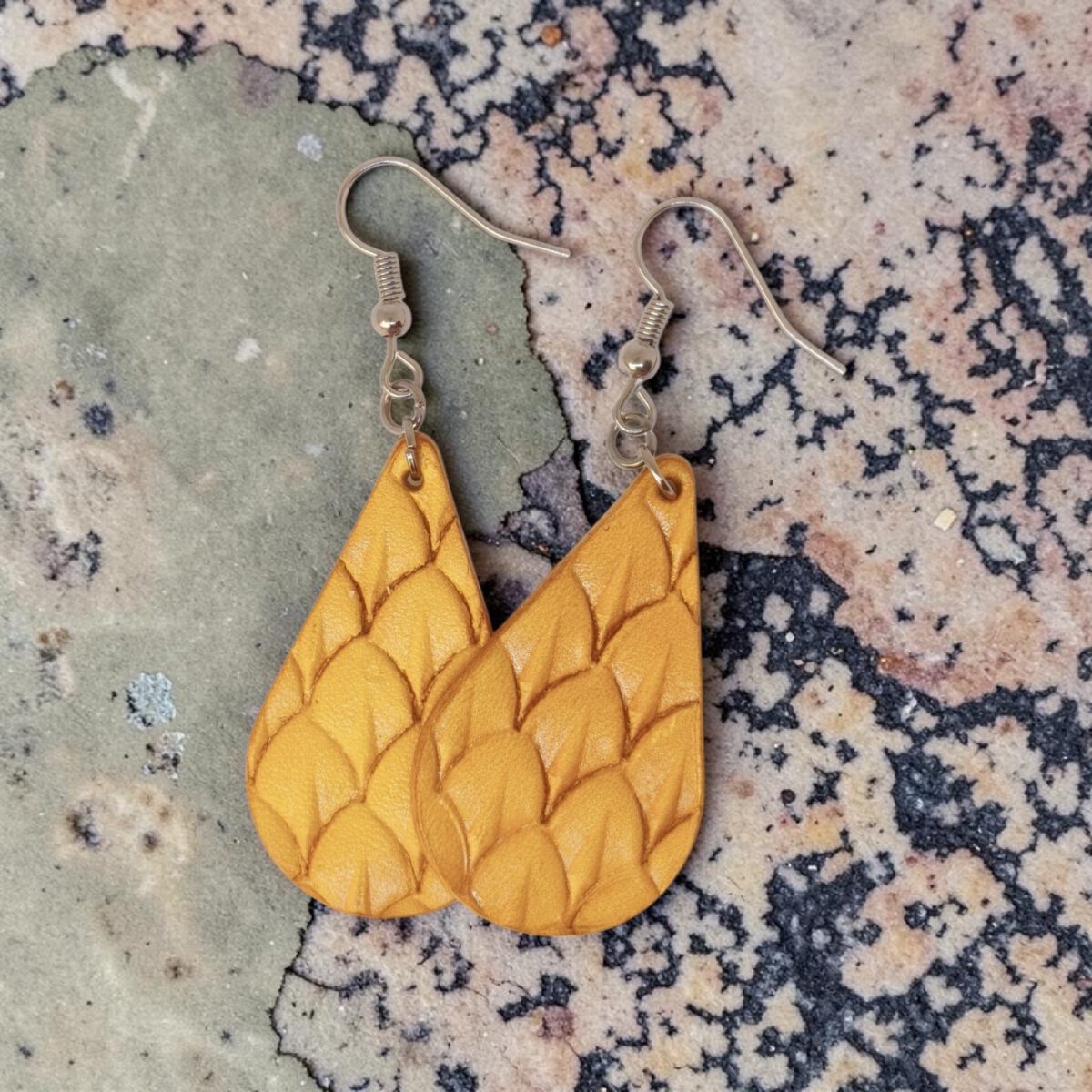 Boucles d'oreilles forme goutte d'eau en cuir teinture jaune, estampages au matoir écailles de poisson, vues de face se chevauchant, suspendues à des boucles argentées sur marbre