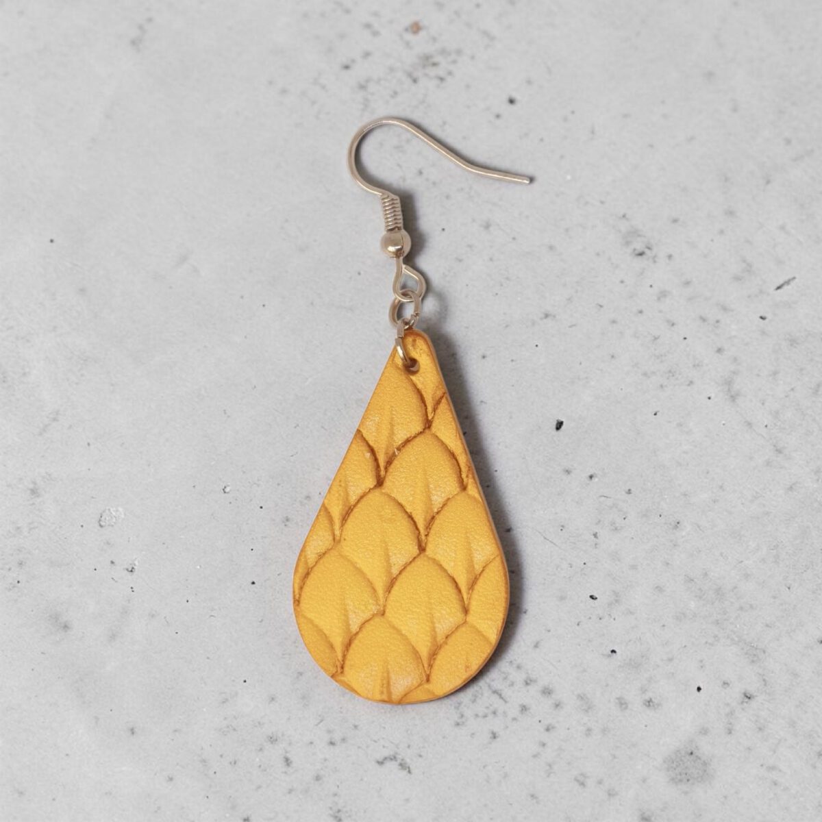 Boucle d'oreille forme goutte d'eau en cuir teinture jaune, estampage au matoir écailles de poisson, vue de face, suspendue à une boucle argentée, sur béton