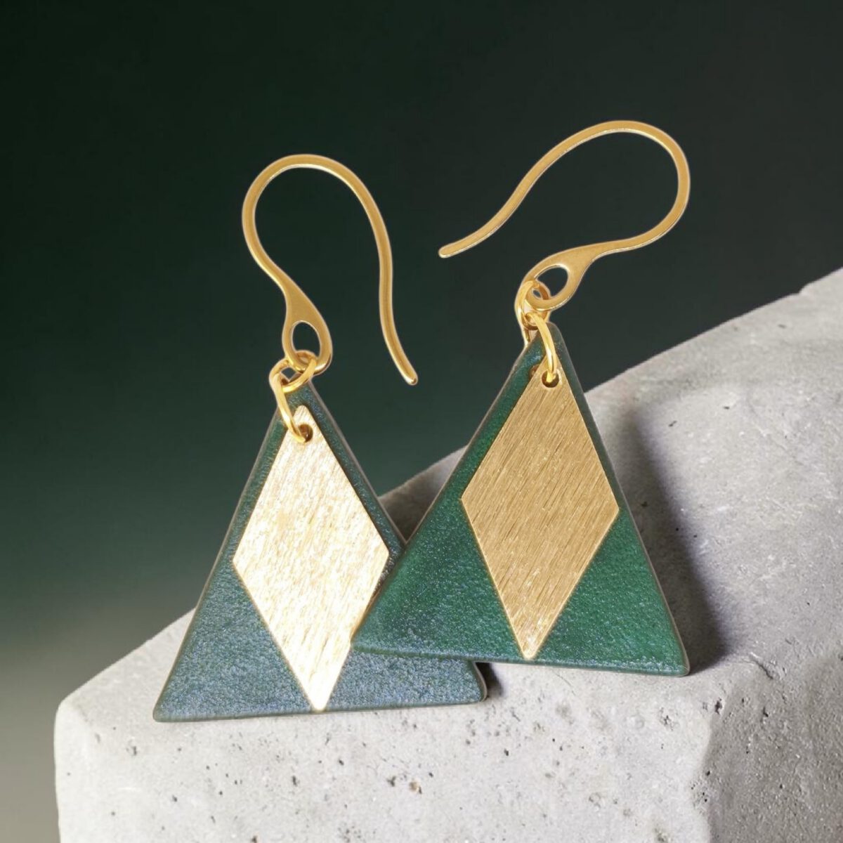 Boucles d'oreilles forme triangulaire en cuir teinture vert sombre, finition lisse et pendants losange dorés brossés vues de face se chevauchant, suspendues à des boucles dorées sur pierre