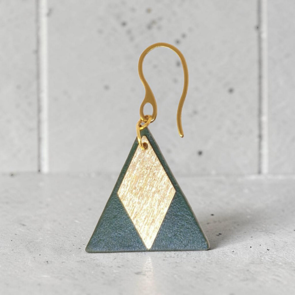 Boucle d'oreille forme triangulaire en cuir teinture vert sombre, finition lisse et pendant losange doré brossé, vue de face, suspendue à une boucle dorée, sur béton