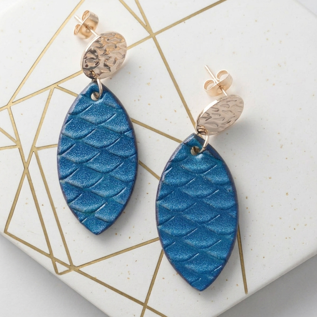 Boucles d'oreilles forme poisson en cuir teinture bleu, texturage poisson large, vues de face, suspendues à des boucles argentées sur fond design doré