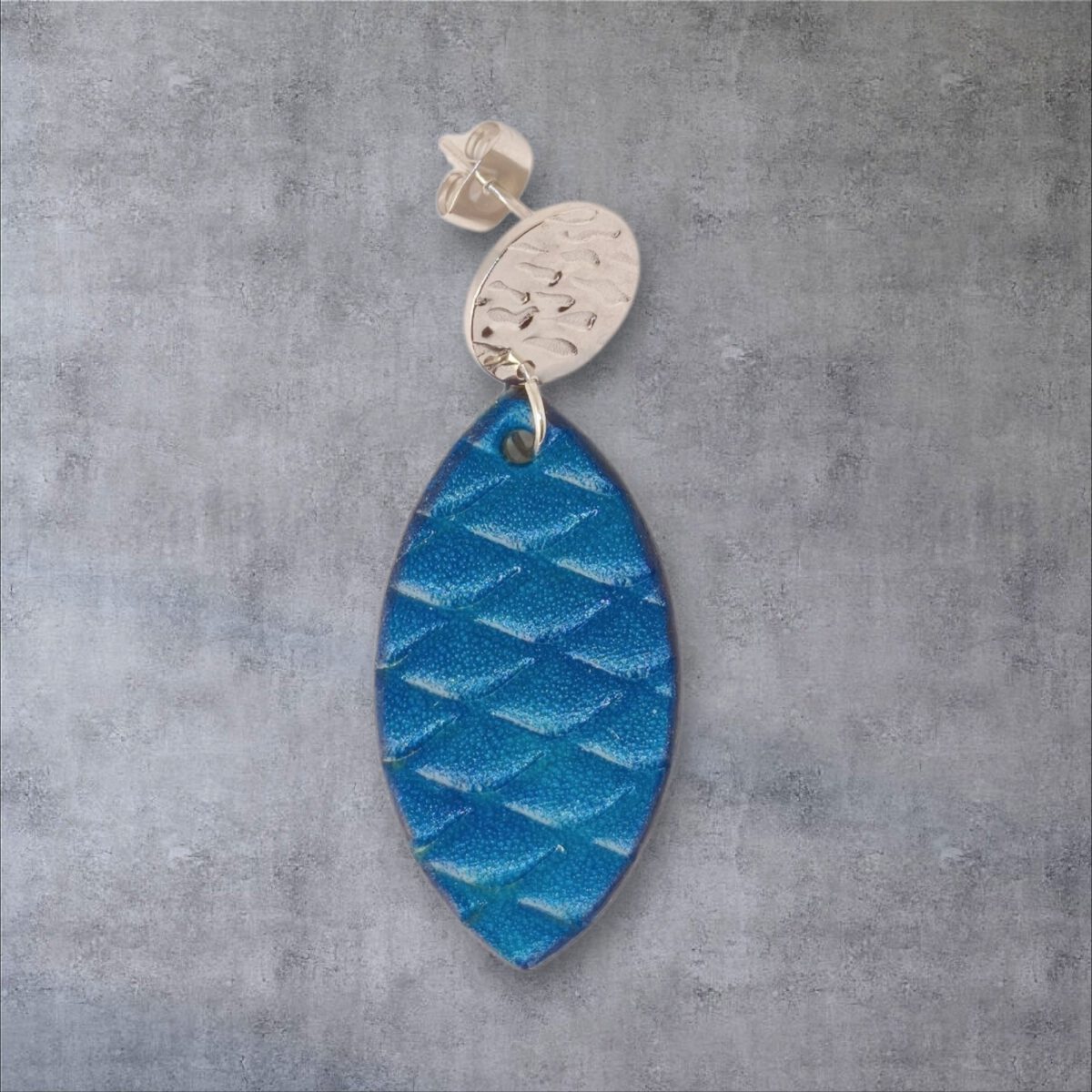 Boucle d'oreille forme poisson en cuir teinture bleu, texturage poisson large, vue de face, suspendue à une boucle argentée, fond gris