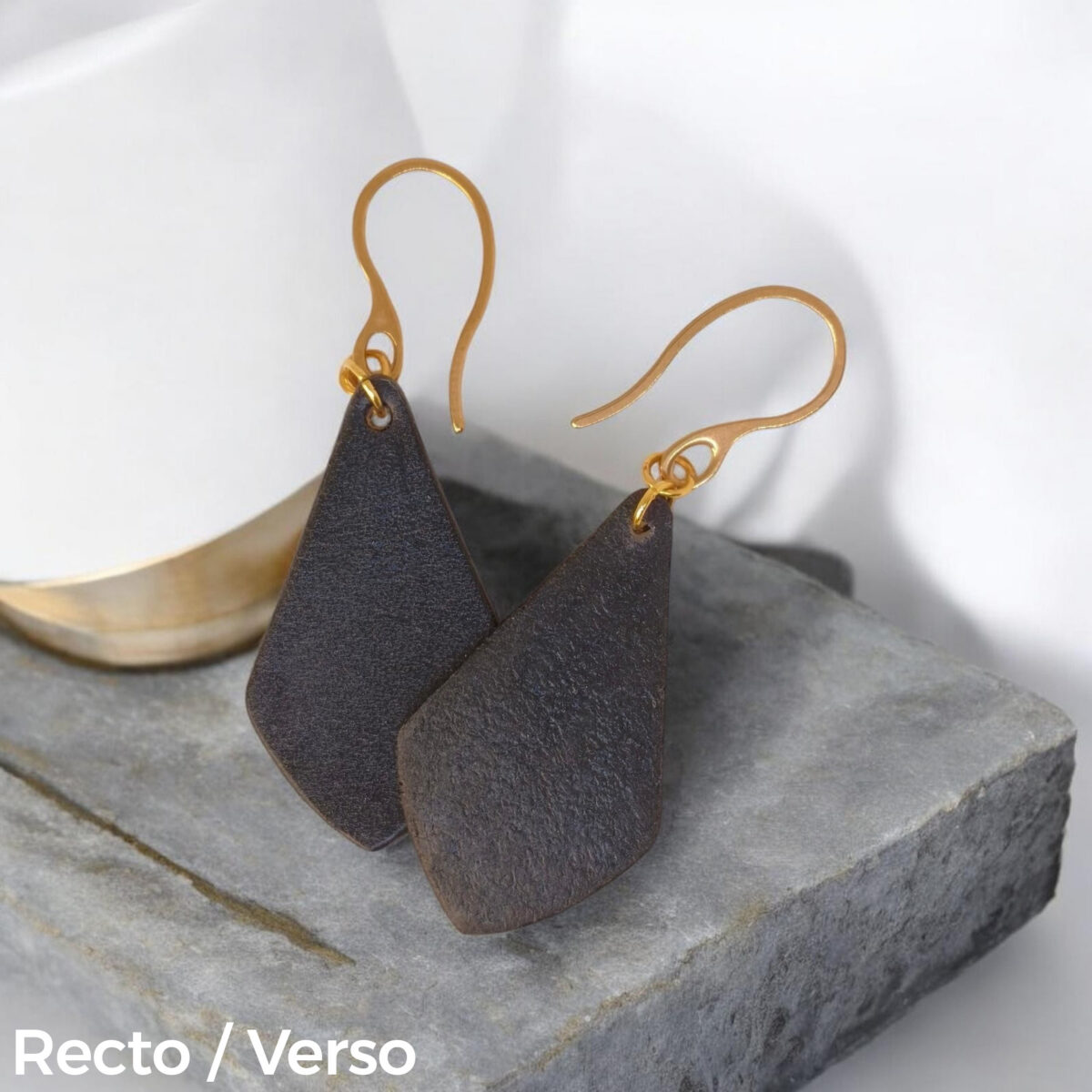 Boucles d'oreilles forme larme en cuir teinture noir charbon, finition lisse, vues recto - verso se chevauchant, suspendues à des boucles dorées sur pierre