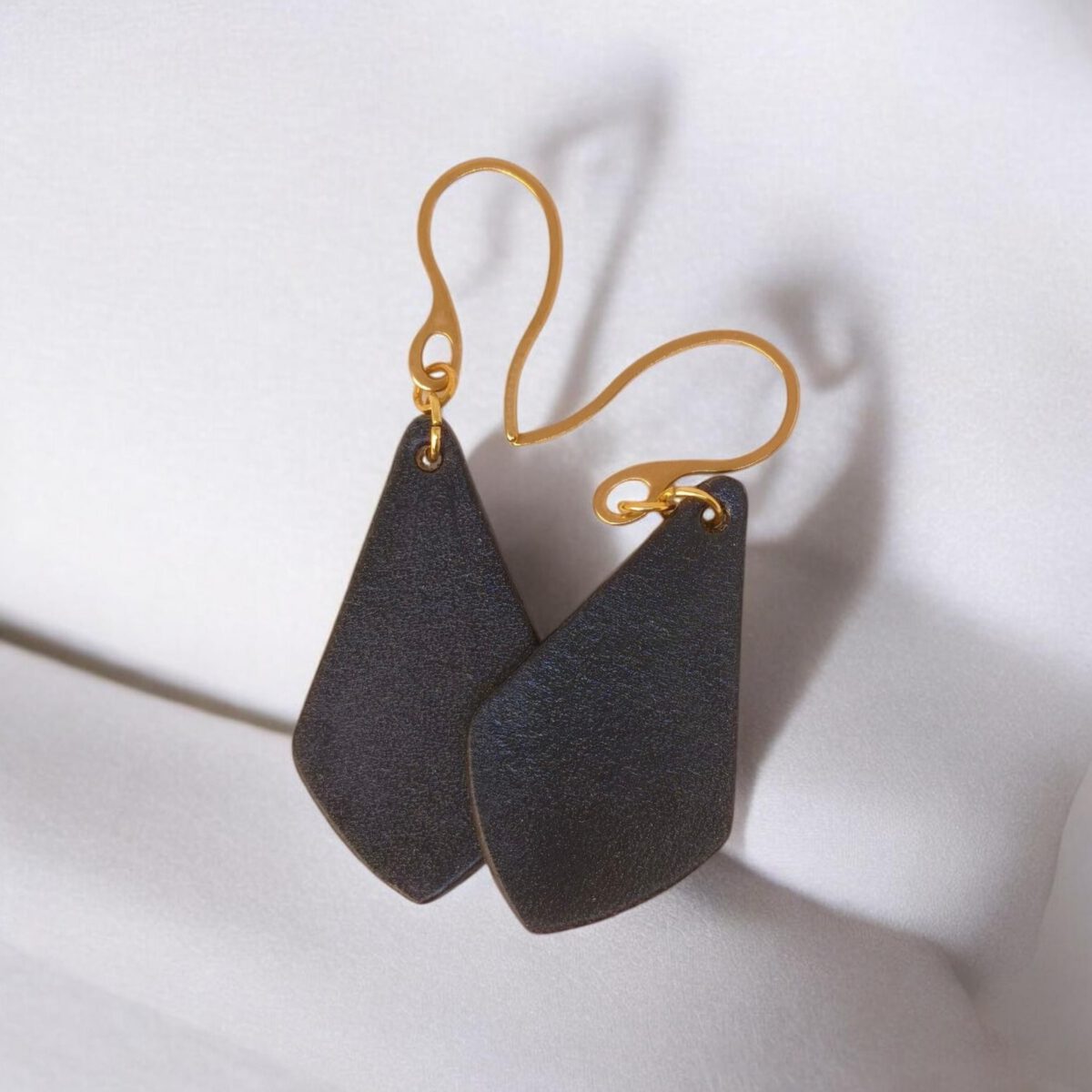 Boucles d'oreilles forme larme en cuir teinture noir charbon, finition lisse, vues de face se chevauchant, suspendues à des boucles dorées sur coussin