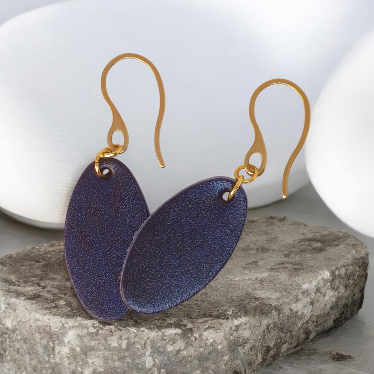 Boucles d'oreilles ovales en cuir sauvage bleu, finition aniline, vues de face se chevauchant, suspendues à des boucles dorées sur pierre