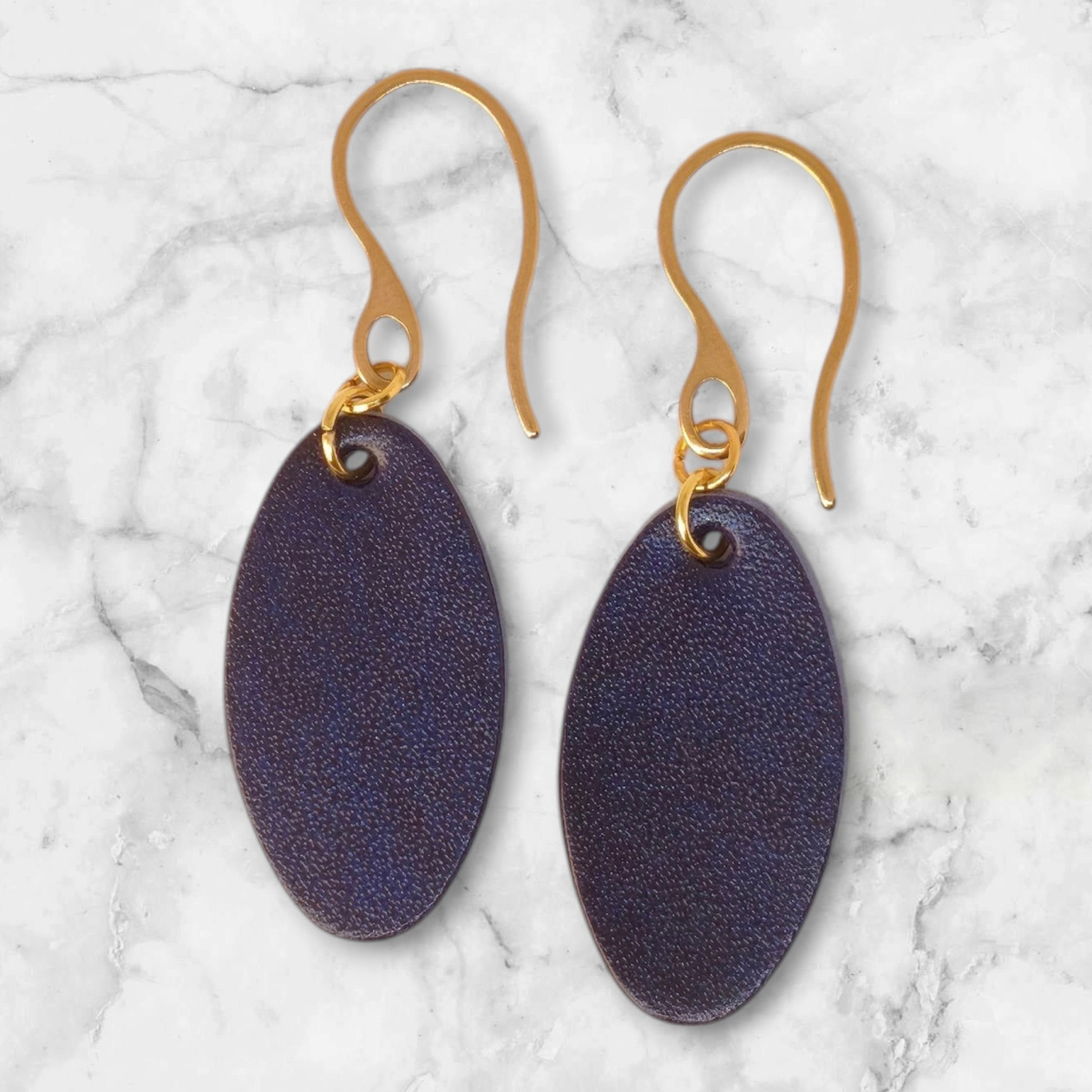 Boucles d'oreilles ovales en cuir sauvage bleu, finition aniline, vues de face, suspendues à des boucles dorées sur marbre