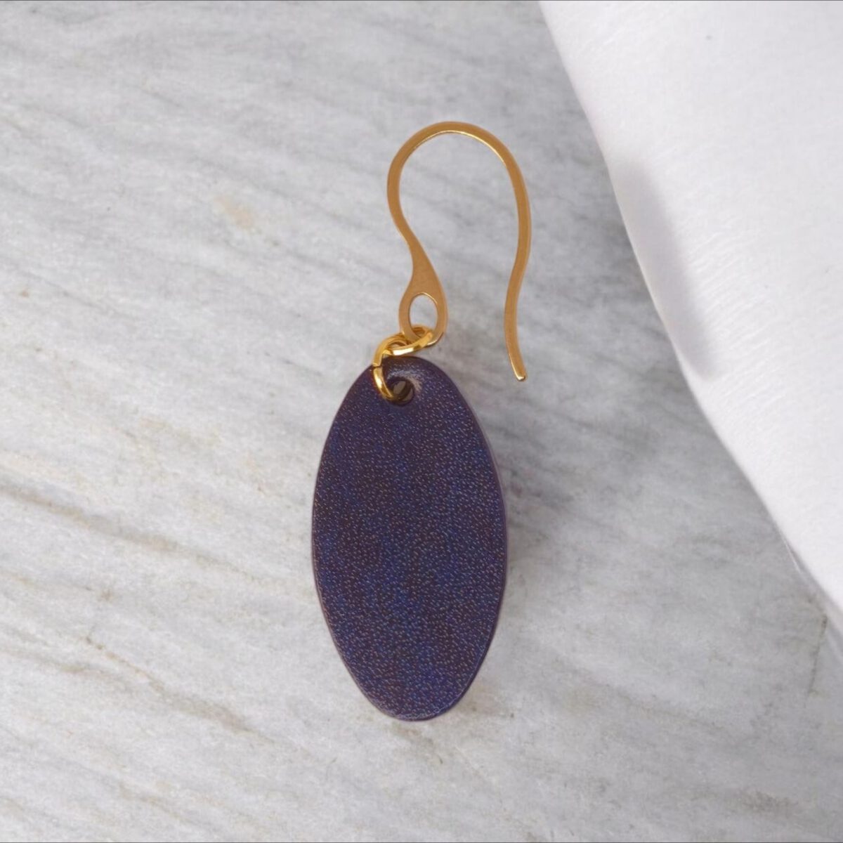 Boucle d'oreille ovale en cuir sauvage bleu, finition aniline, vue de face, suspendue à une boucle dorée, fond design