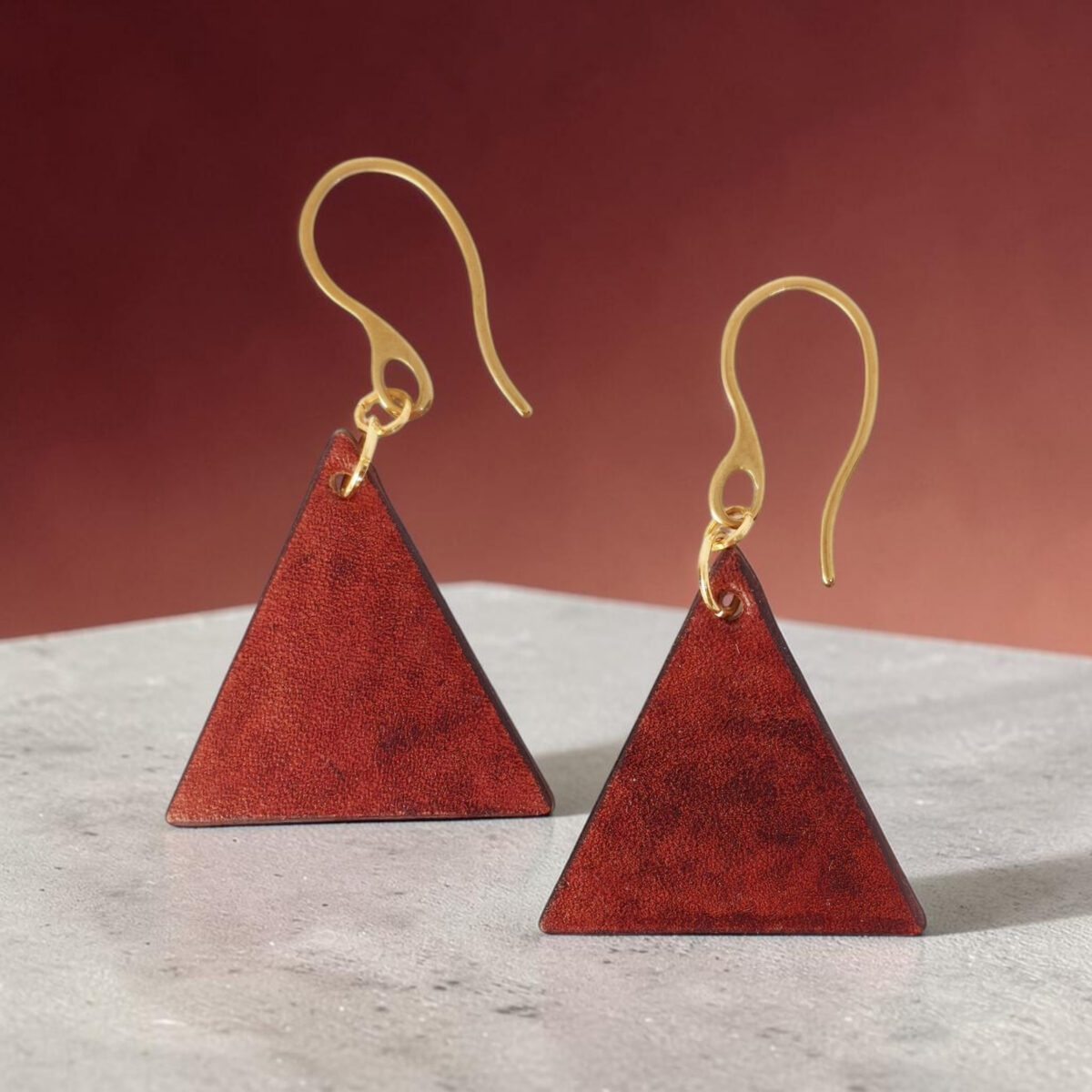 Boucles d'oreilles forme triangulaire en cuir teinture cognac, finition lisse, vues de face, suspendues à des boucles dorées sur béton