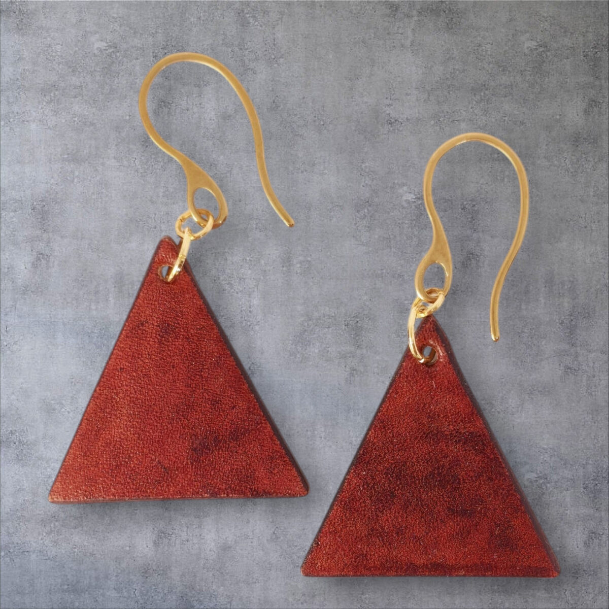 Boucles d'oreilles forme triangulaire en cuir teinture cognac, finition lisse, vues de face, suspendues à des boucles dorées sur fond gris