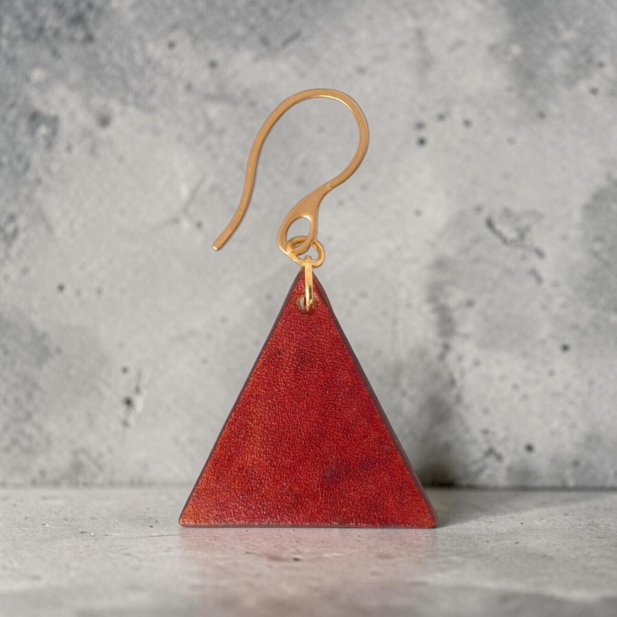 Boucle d'oreille forme triangulaire en cuir teinture cognac, finition lisse, vue de face, suspendue à une boucle dorée, sur béton ciré