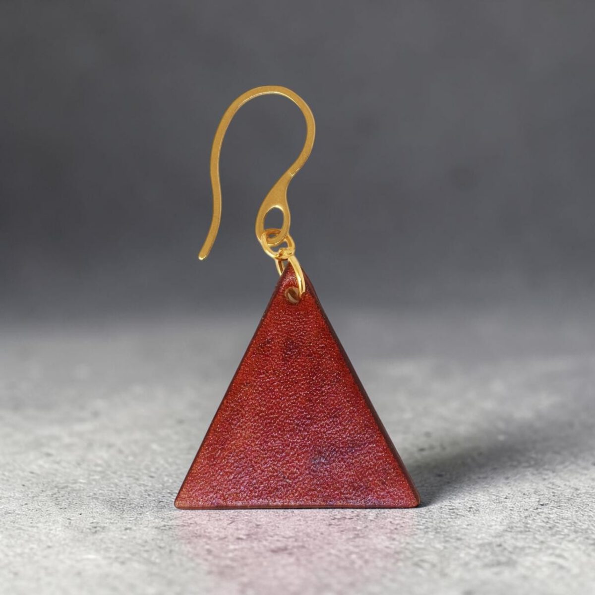 Boucle d'oreille forme triangulaire en cuir teinture cognac, finition lisse, vue de face, suspendue à une boucle dorée, sur béton