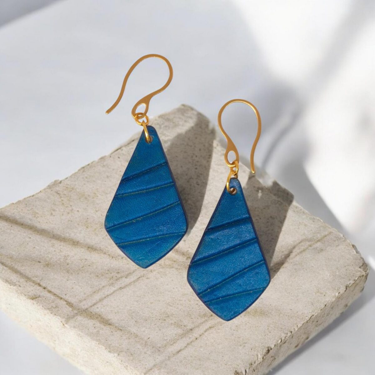 Boucles d'oreilles forme larme en cuir teinté bleu, finition galet, vues de face, suspendues à des boucles dorées, sur pierre