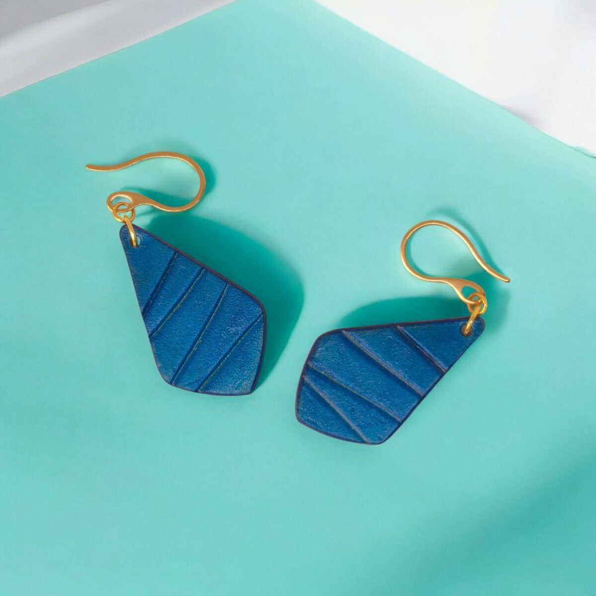 Boucles d'oreilles forme larme en cuir teinté bleu, finition galet, vues de face, suspendues à des boucles dorées sur fond design
