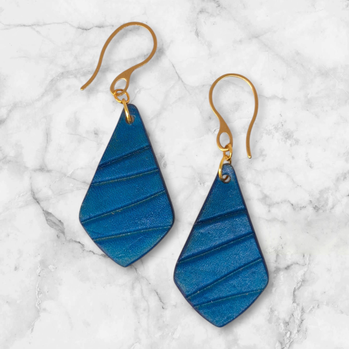 Boucles d'oreilles forme larme en cuir teinté bleu, finition galet, vues de face, suspendues à des boucles dorées sur marbre