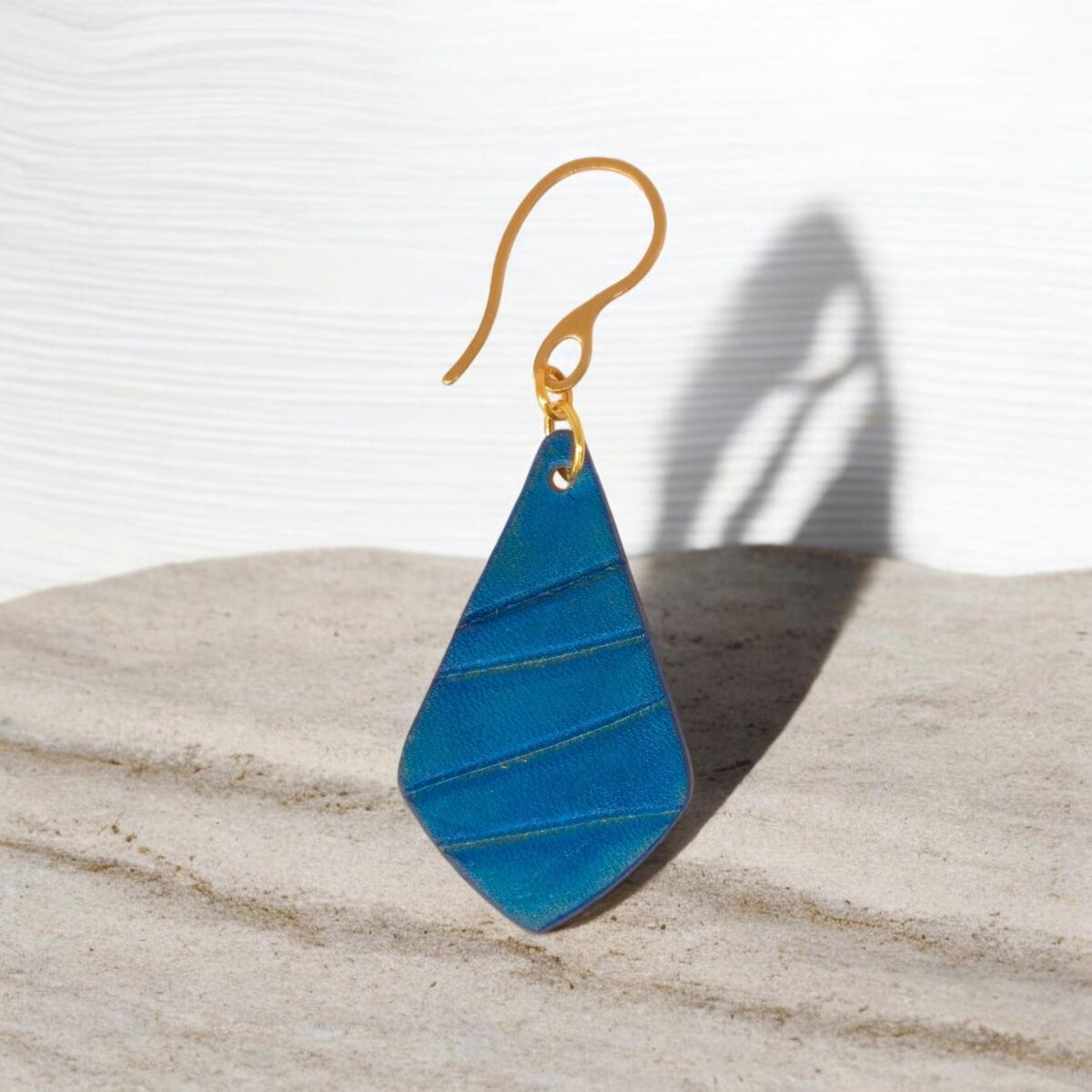 Boucle d'oreille forme larme en cuir teinté bleu, finition galet, vue de face, suspendue à une boucle dorée, fond pierre
