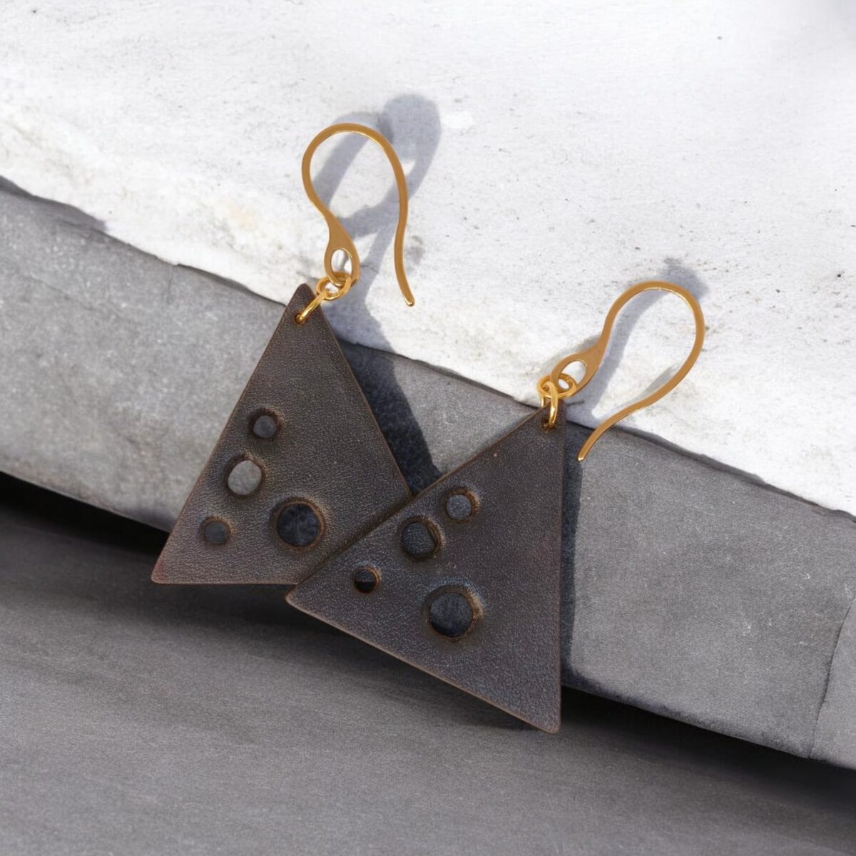 Boucles d'oreilles forme triangulaire en cuir teinture noire, finition lisse et ses trous, vues de face se chevauchant, suspendues à des boucles dorées