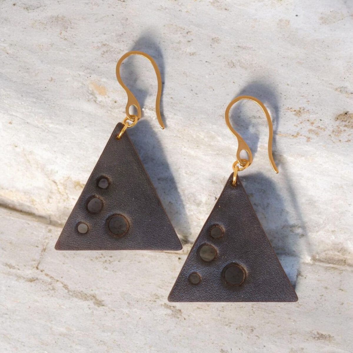 Boucles d'oreilles forme triangulaire en cuir teinture noire, finition lisse et ses trous, vues de face, suspendues à des boucles dorées sur pierre