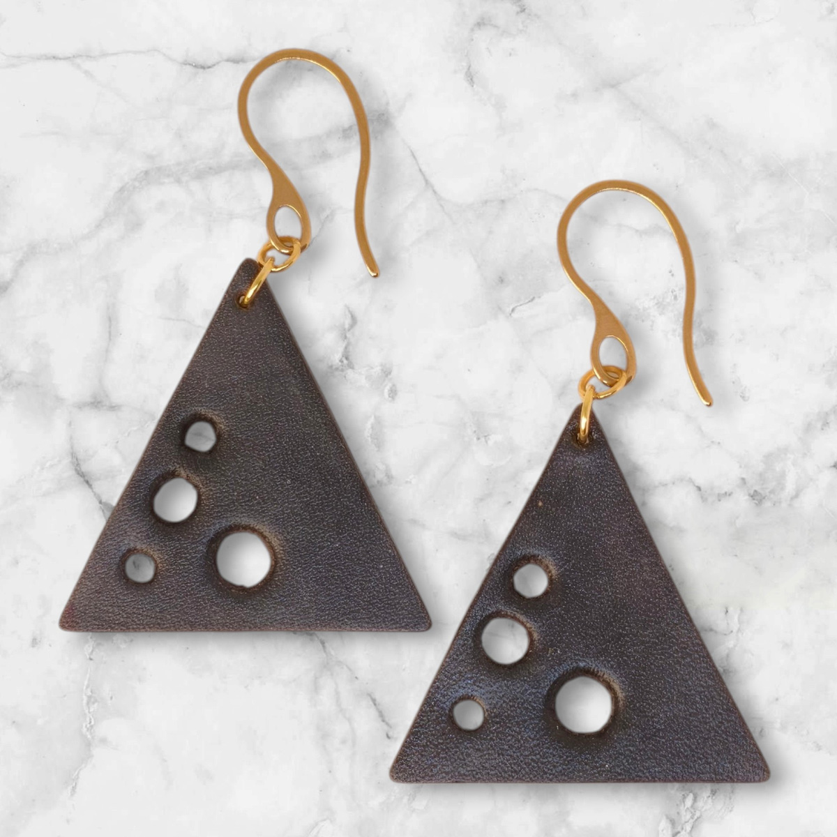 Boucles d'oreilles forme triangulaire en cuir teinture noire, finition lisse et ses trous, vues de face, suspendues à des boucles dorées sur marbre